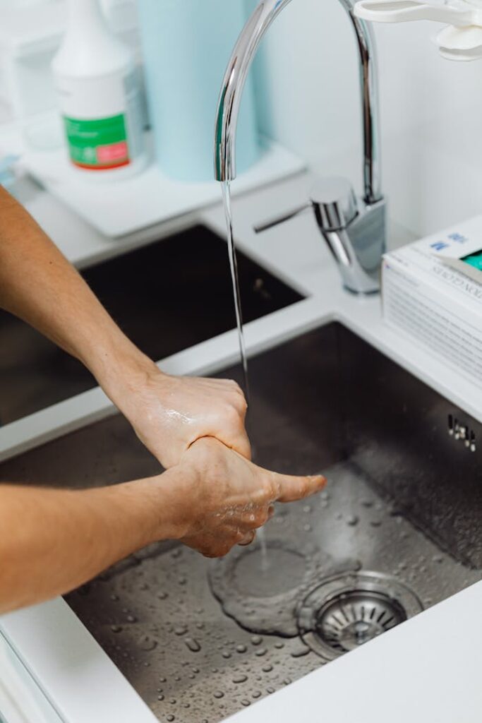 pexels-photo-6627718 Hands being washed under running water in a modern stainless steel sink, promoting hygiene.